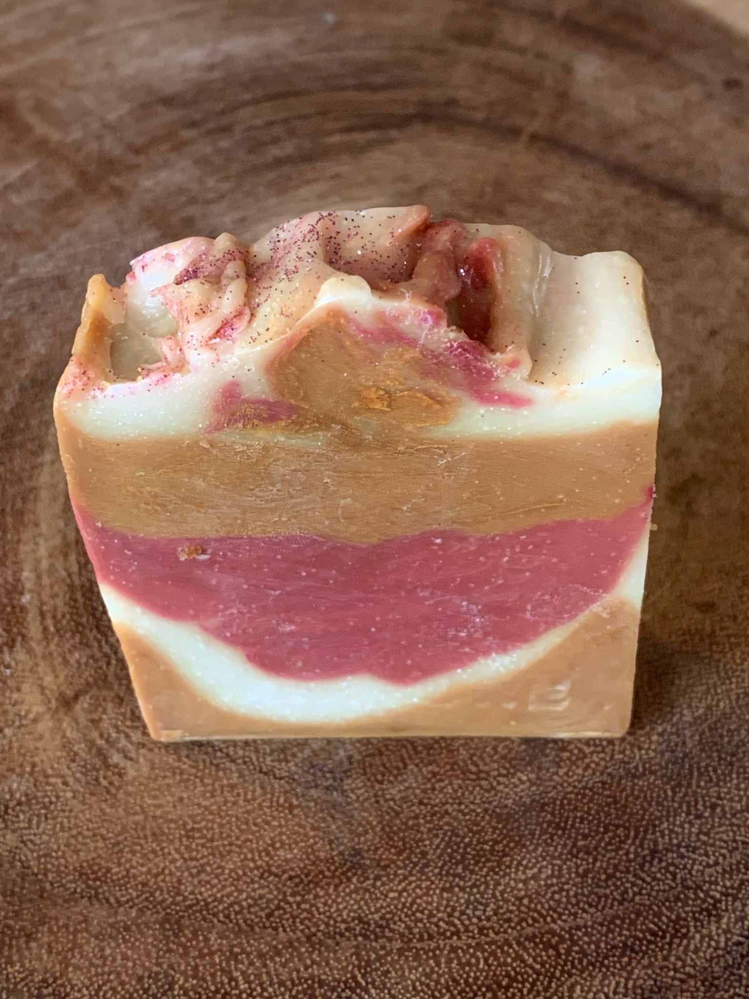 A bar of soap with layers of beige and pink, topped with a sprinkle of pink powder, on a wooden surface.