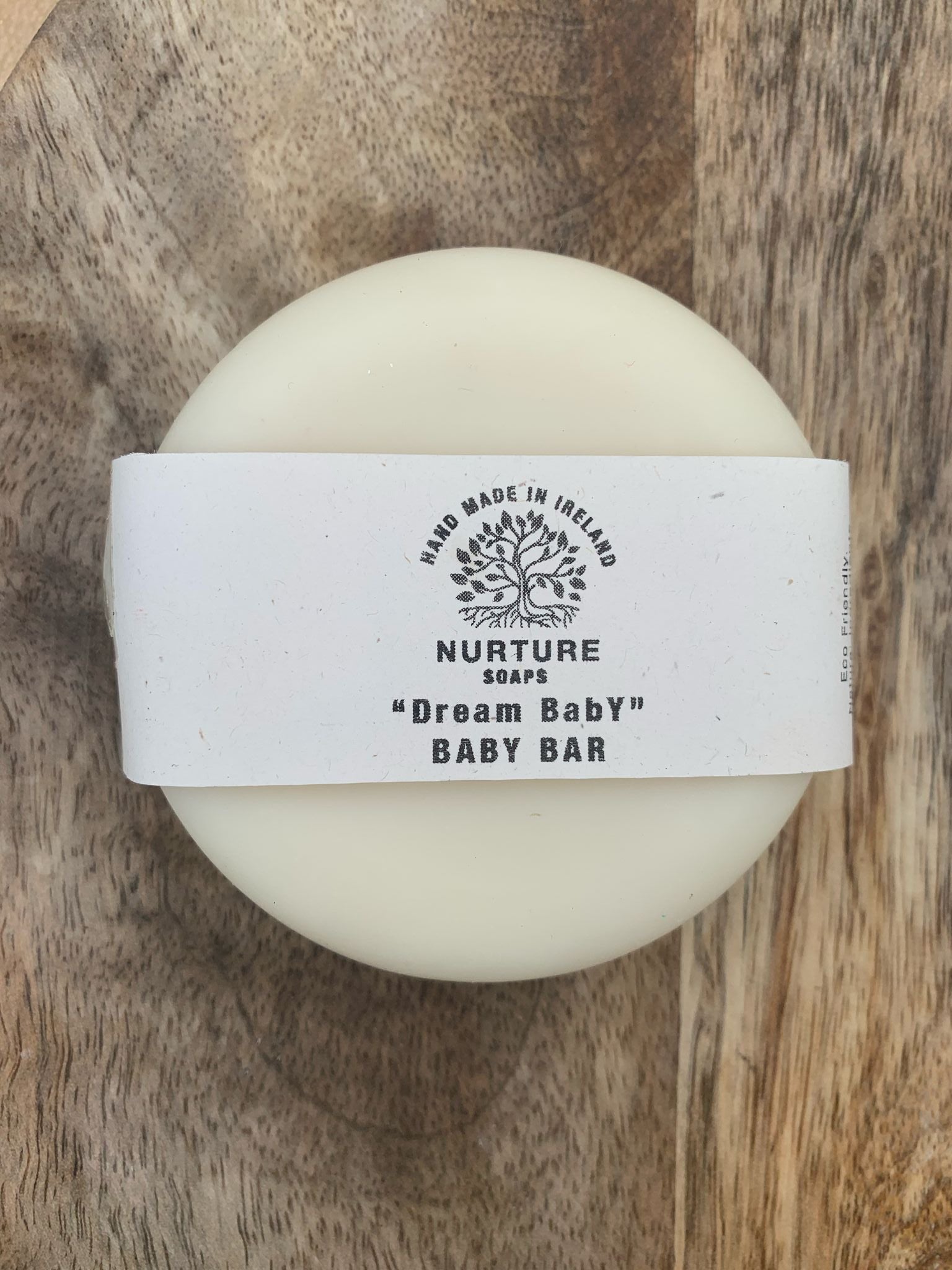 A round bar of soap with a label that reads 'NURTURE SOAPS Dream Baby BABY BAR'. The soap is placed on a wooden surface.