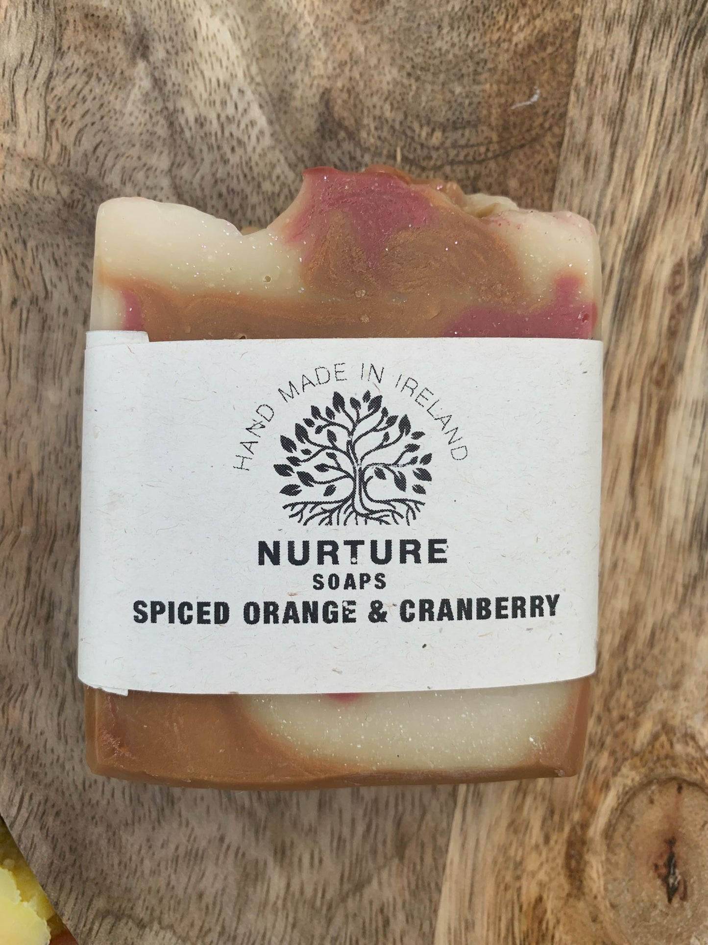 A soap bar with a swirl pattern, primarily beige with red accents, labeled 'NURTURE SOAPS SPICED ORANGE & CRANBERRY'.