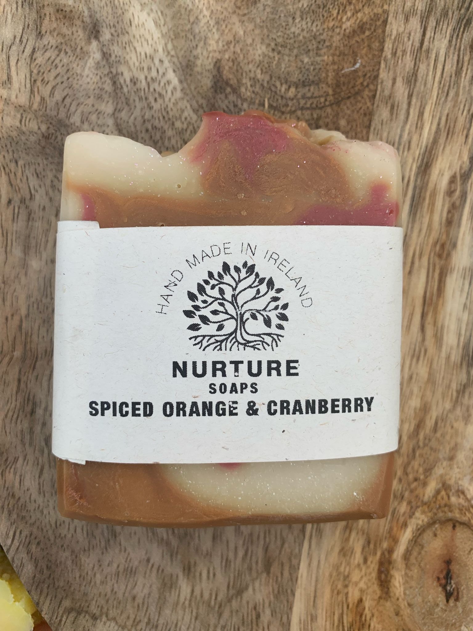 A soap bar with a swirl pattern, primarily beige with red accents, labeled 'NURTURE SOAPS SPICED ORANGE & CRANBERRY'.