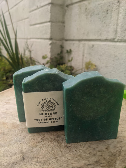 Three bars of soap with a dark green color, placed on a surface with a white wall and greenery in the background.