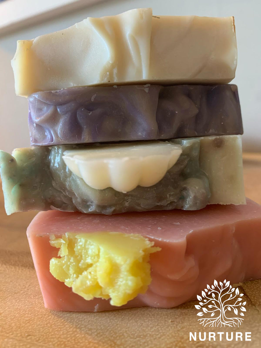 A stack of three bars of soap with varying shades of purple, green, and pink, with the Nurture logo on the bottom right.
