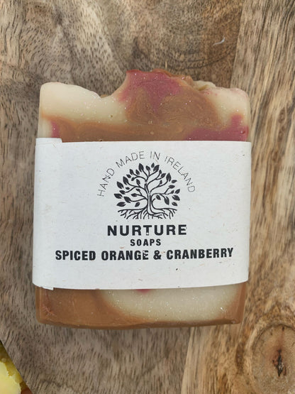 A soap bar with a swirl pattern, primarily beige with red accents, labeled 'NURTURE SOAPS SPICED ORANGE & CRANBERRY'.